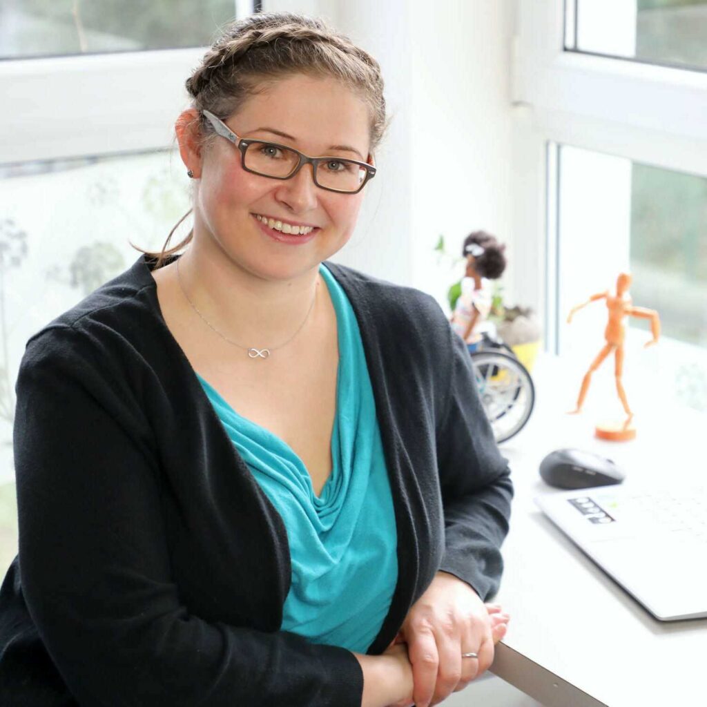 Martina Götschel sitzt lächelnd an einem Schreibtisch vor einem Laptop, hinter ihr erkennt man eine Rollstuhl-Barbie und ein Bewegungs-Männchen.